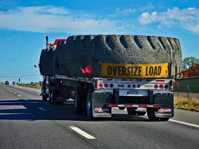 A truck on a road carrying a giant tire with a banner pinned to it that says 'oversize load'