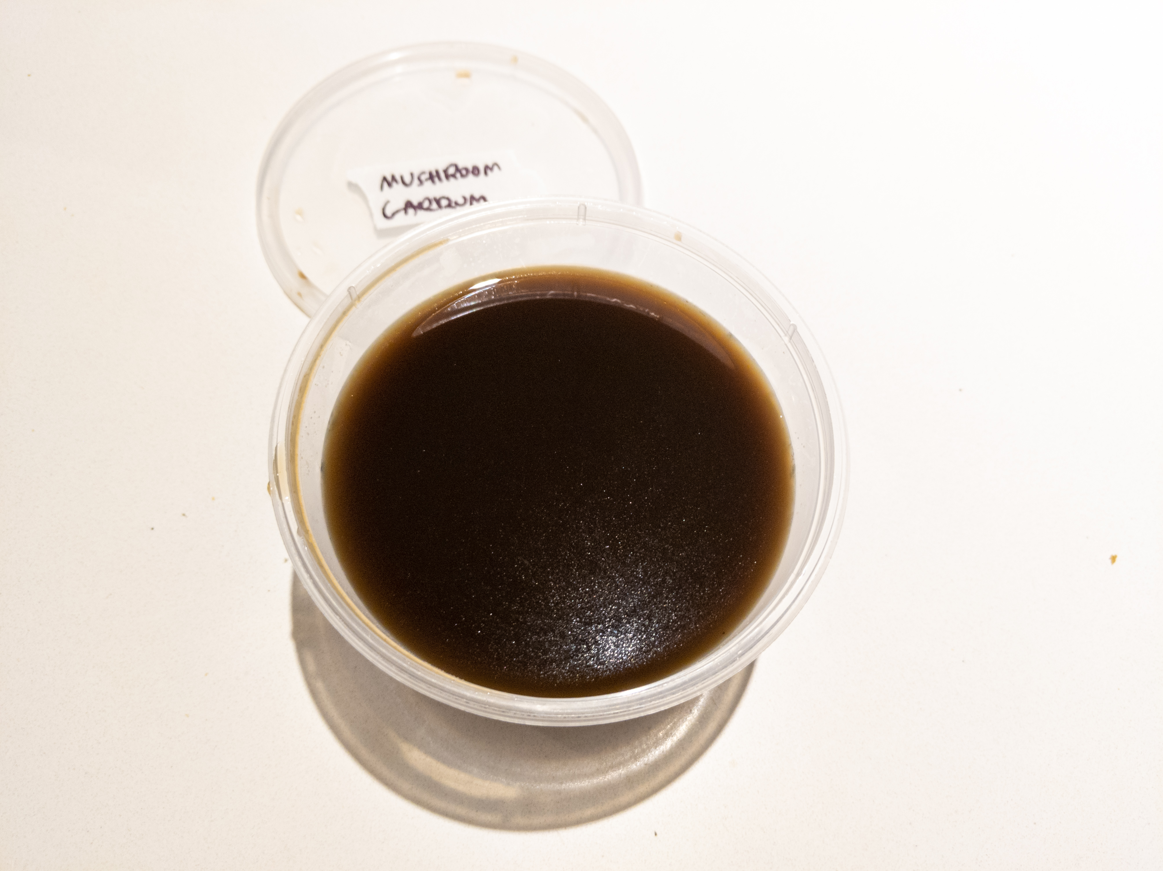 A plastic deli container filled with a brown liquid. The container is on a white counter, and there is a deli lid behind it labeled 'mushroom garrum'