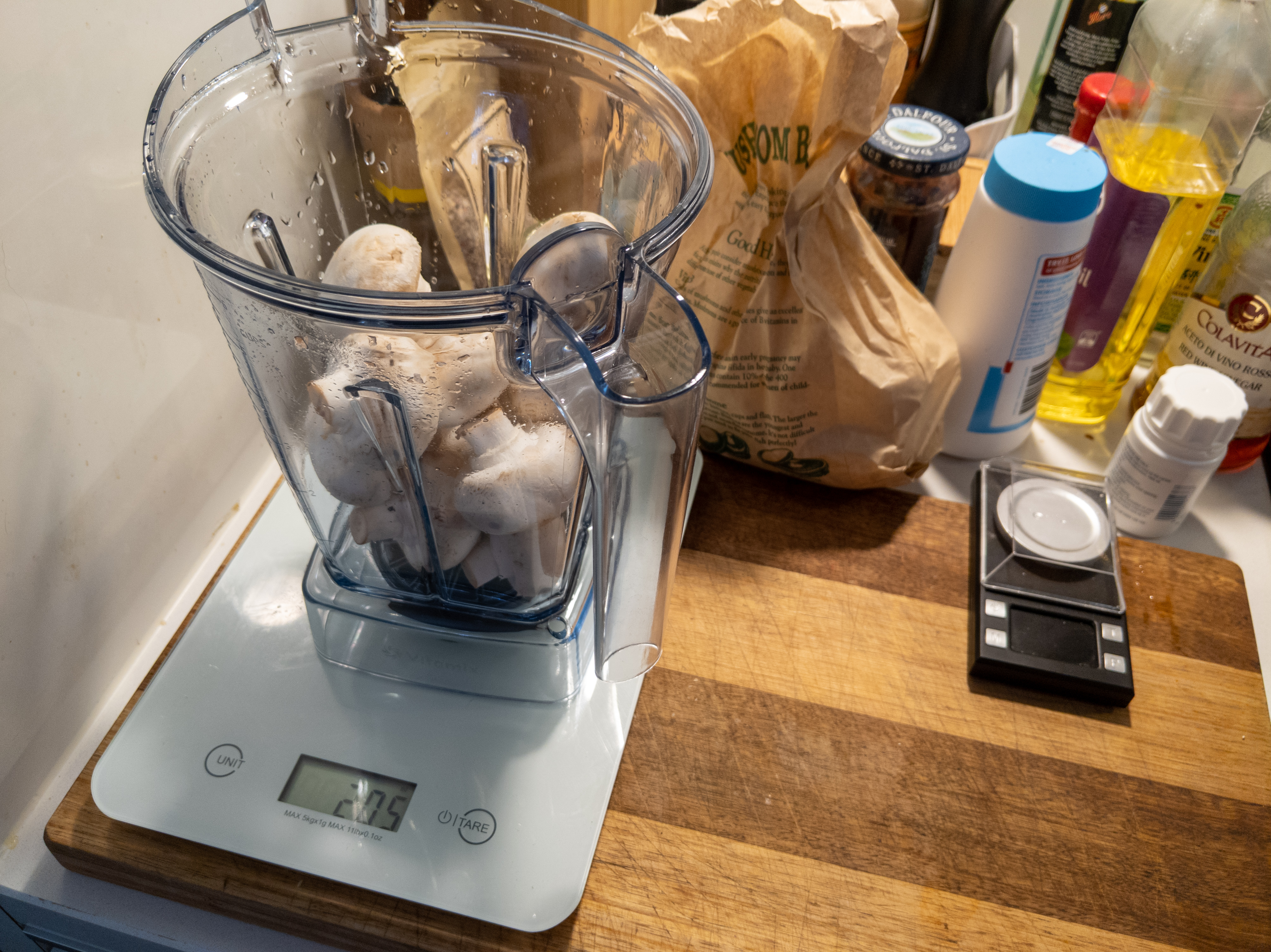 A blender canister on a scale, filled with mushrooms. The scale is sitting on a wooden cutting board on a cluttered kitchen counter.
