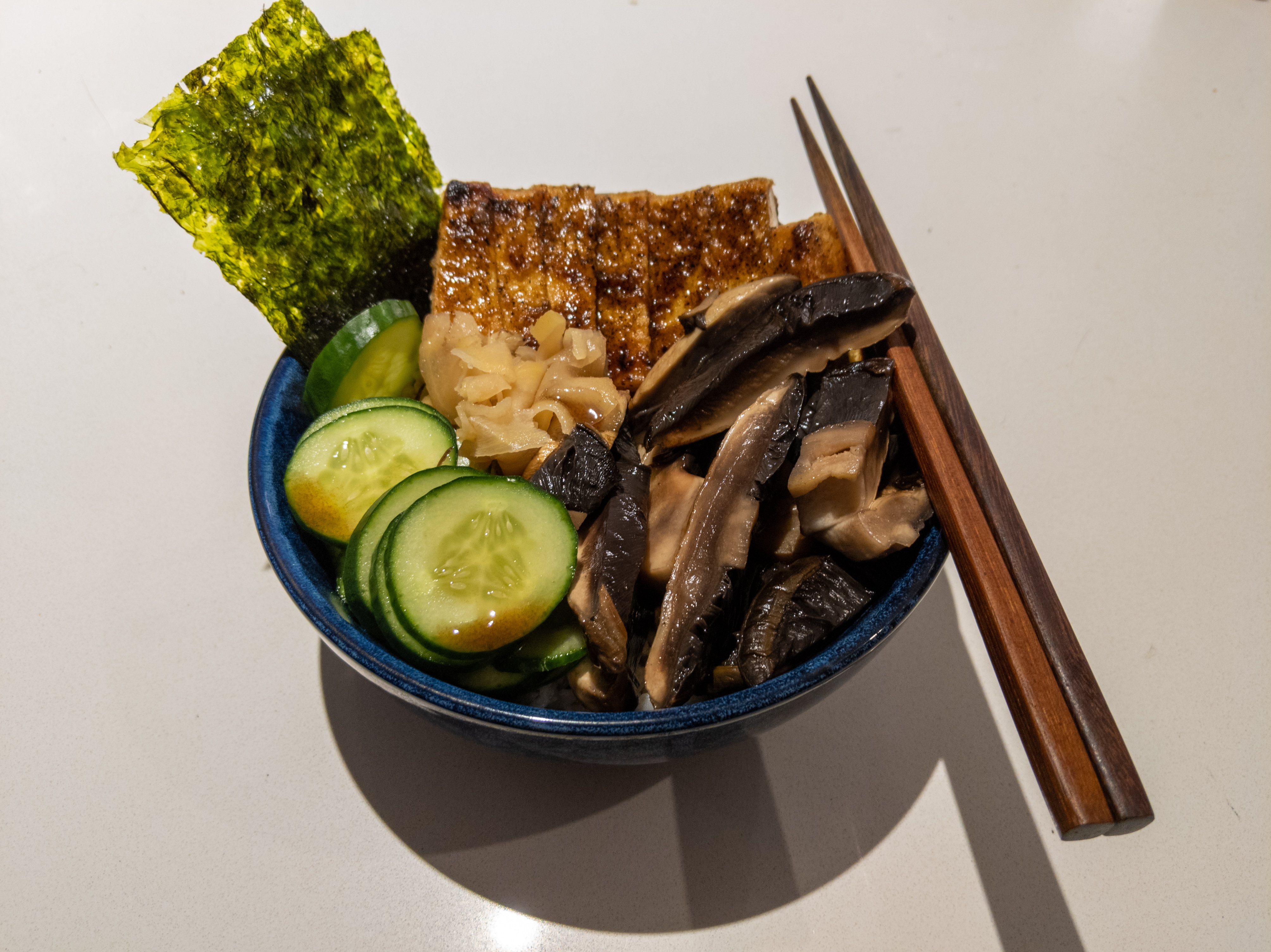 A blue bowl with some chopsticks on the side, filled with rice and toppings. The toppings are pickled ginger, wheels of cucumber, slices of mushroom, slices of tofu, and some pieces of seaweed.