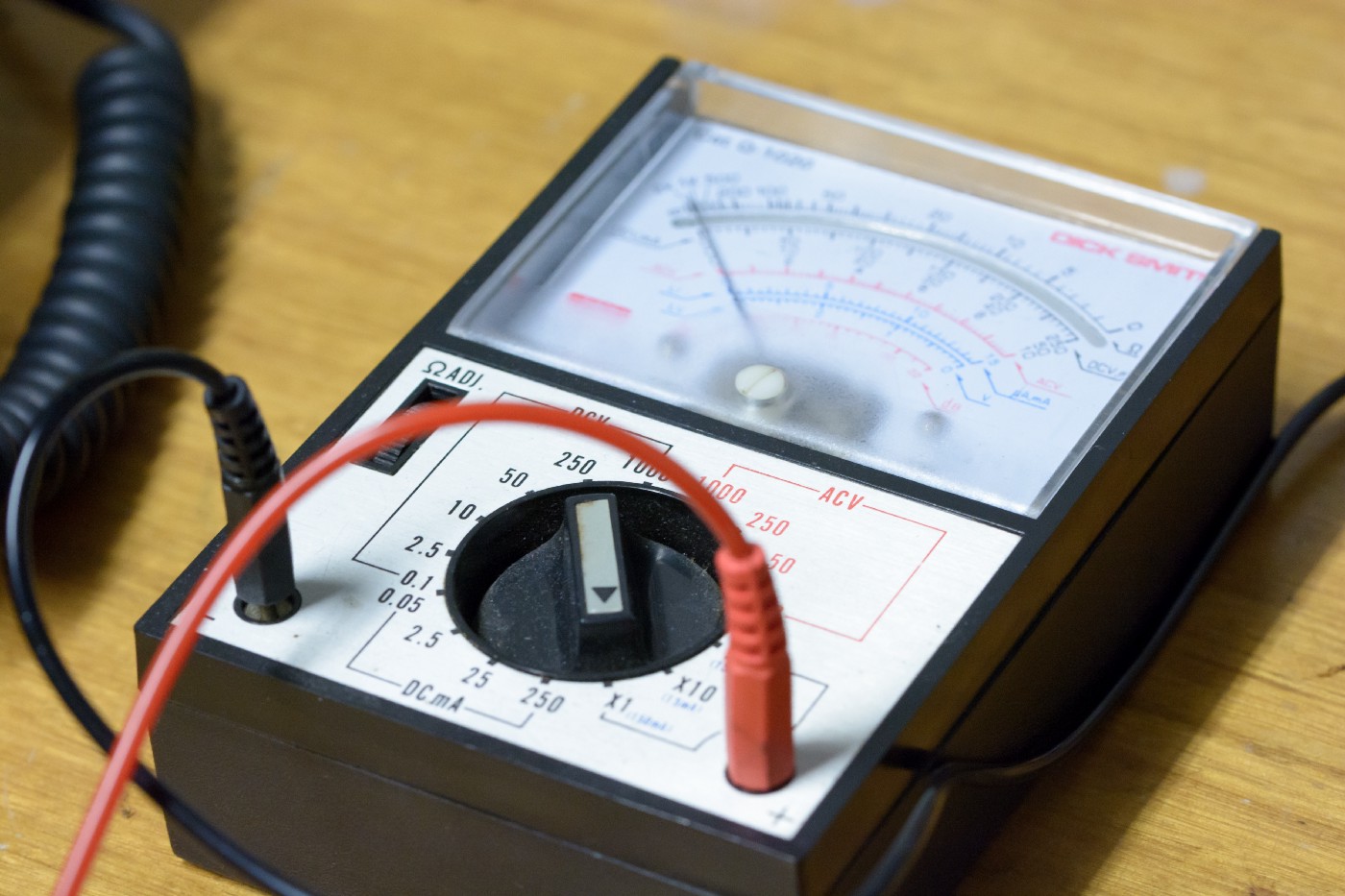 A close up image of an analog multimeter, with a red and a black test lead emerging from two ports.