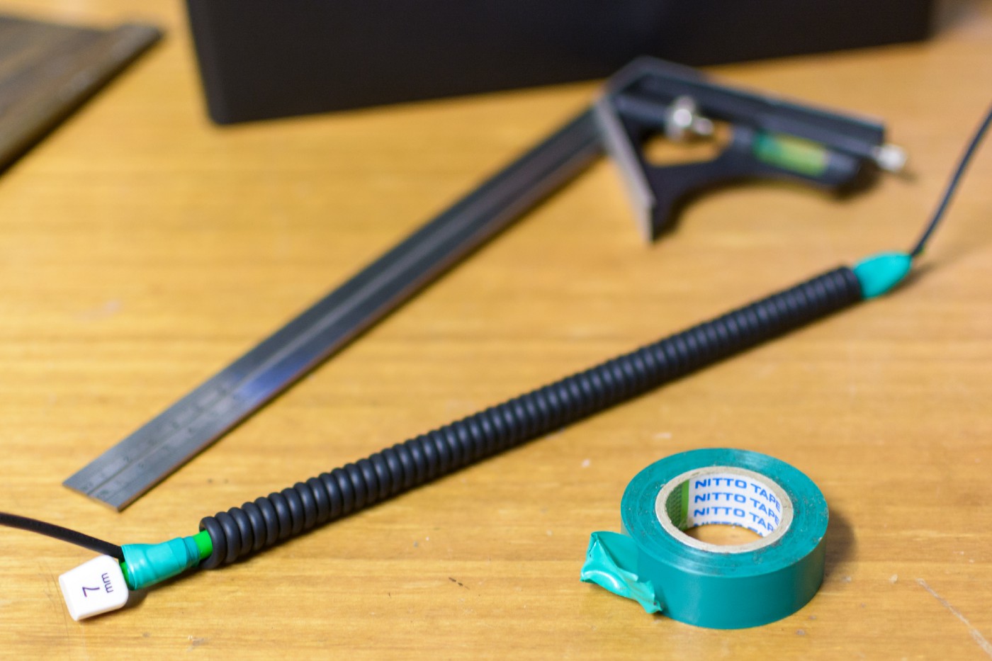 An image of a black cable coiled around a knitting needle, resting on a wooden bench top. There is a roll of green electrical tape sitting nearby