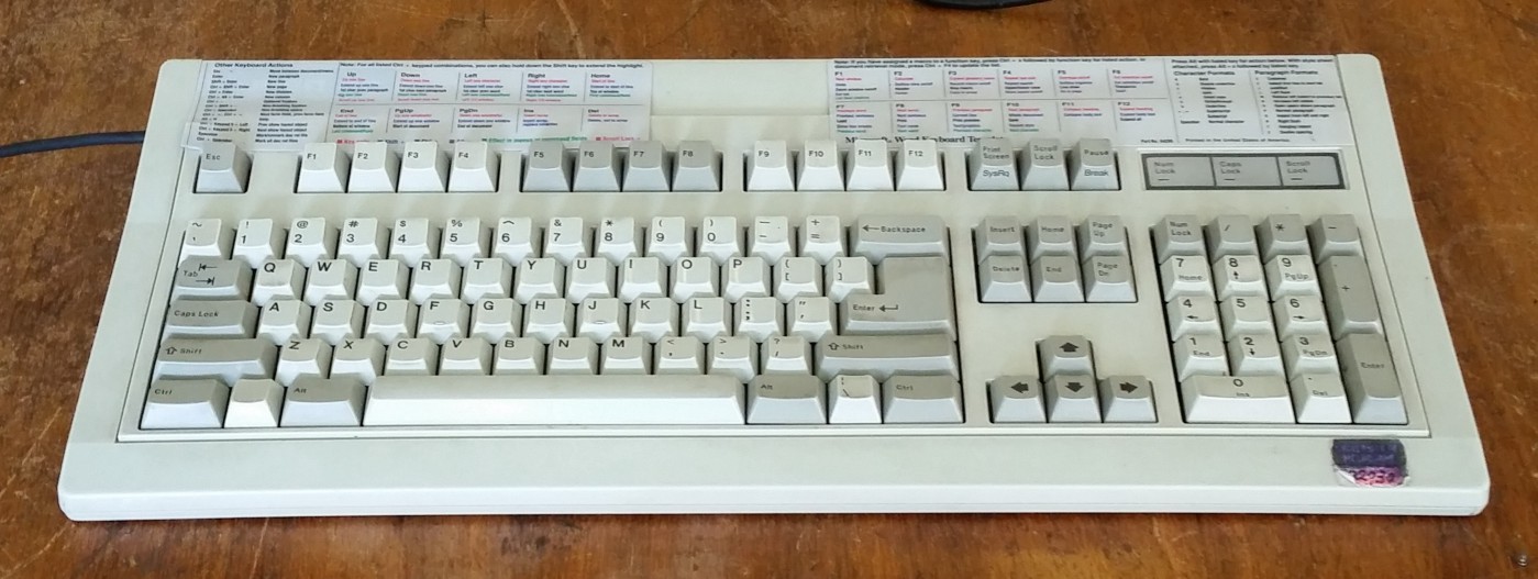 An image of a large, old keyboard. It has keyboard shortcuts for a program printed out and taped to the top of it.