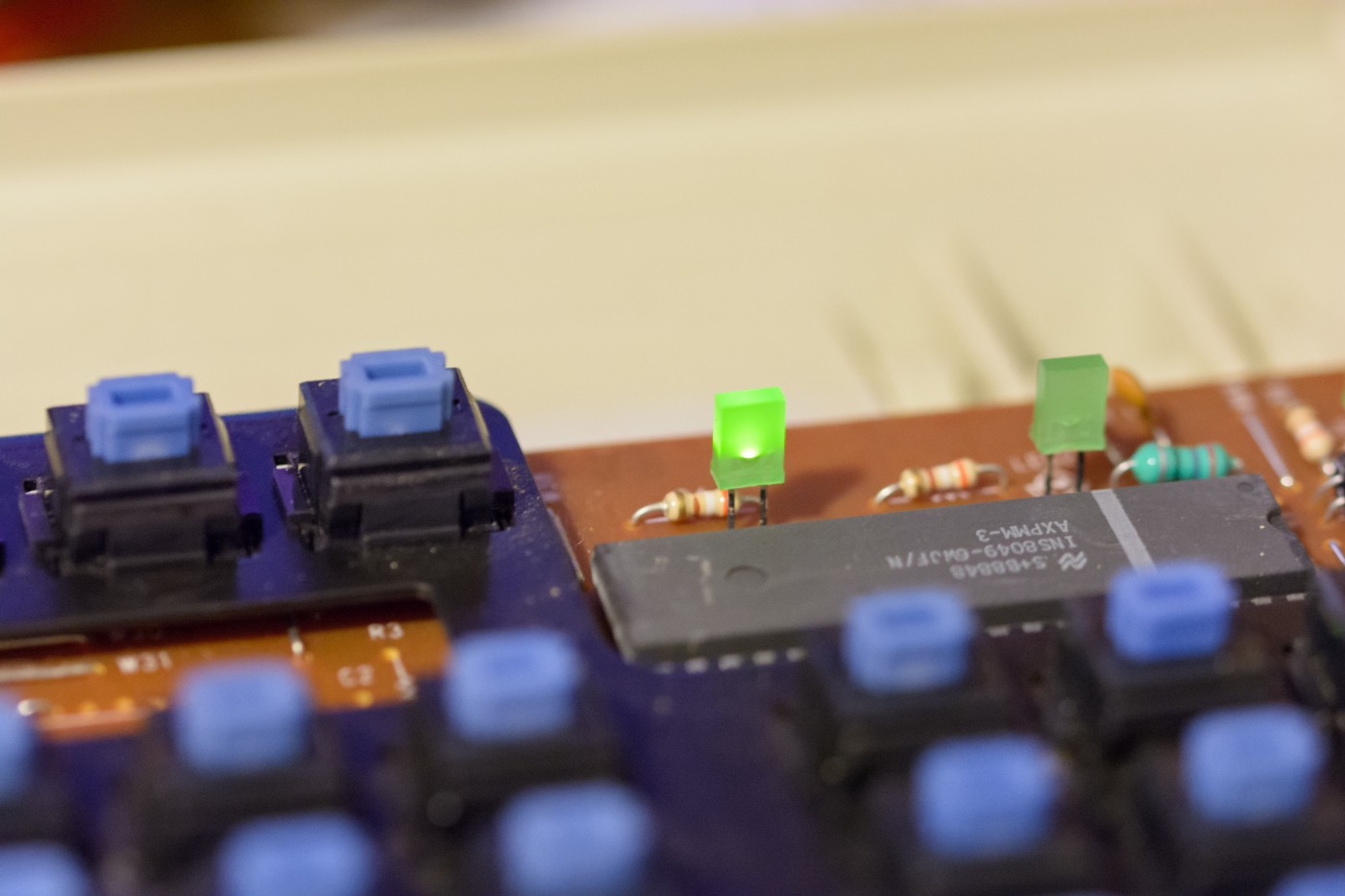 A macro image of the mainboard of a keyboard, centered on a rectangular prism shaped LED. The LED is lit up and green colored.