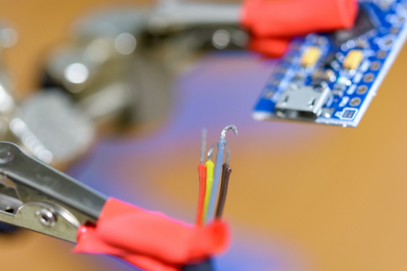 A macro photo of four wires that have been tinned with solder and hooked, being held in a third-hand. In the background is a microcontroller on a breakout board.